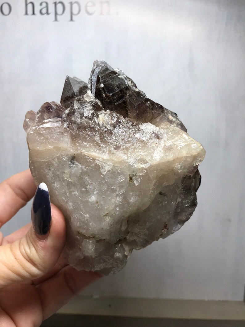 May include: A large, clear quartz crystal cluster with a smoky quartz point on top. The crystal has a natural, rough texture and is a beautiful example of a natural crystal formation.