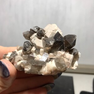 Puede incluir: Un c&uacute;mulo de cristales de cuarzo ahumado con una matriz de color marr&oacute;n claro, casi blanco. Los cristales son transl&uacute;cidos y tienen una apariencia ligeramente nublada.
