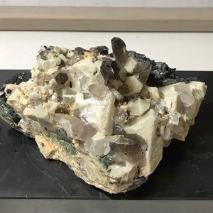 May include: A cluster of smoky quartz crystals embedded in a matrix of white and gray rock. The crystals are translucent and have a smoky brown color. The rock is rough and textured.