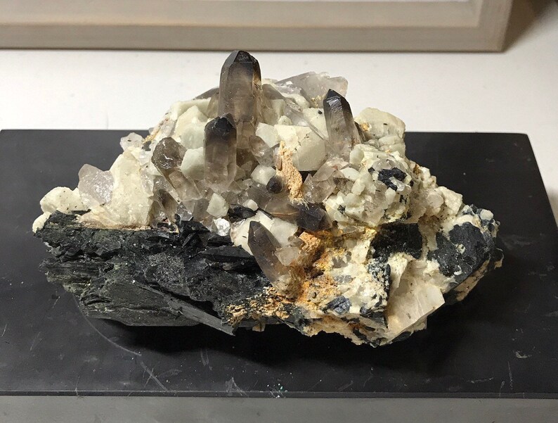 May include: A cluster of smoky quartz crystals on a black matrix. The crystals are clear and have a smoky brown color. The matrix is black and has a rough texture.