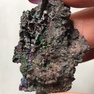 Pu&ograve; includere: Primo piano di un minerale grigio scuro iridescente con una superficie ruvida e testurizzata. Il minerale presenta una variet&agrave; di colori, tra cui viola, verde e blu.