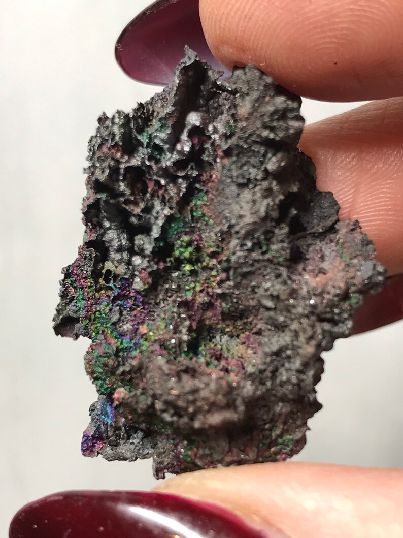 Pu&ograve; includere: Primo piano di un minerale grigio scuro iridescente con una superficie ruvida e testurizzata. Il minerale presenta una variet&agrave; di colori, tra cui viola, verde e blu.