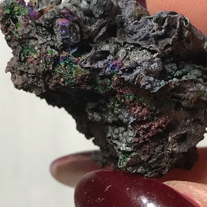 Pu&ograve; includere: Primo piano di un minerale grigio scuro iridescente con una superficie ruvida e testurizzata. Il minerale ha un'apparenza metallica e mostra una variet&agrave; di colori, tra cui verde, blu e viola.
