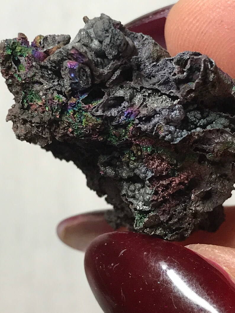 Pu&ograve; includere: Primo piano di un minerale grigio scuro iridescente con una superficie ruvida e testurizzata. Il minerale ha un'apparenza metallica e mostra una variet&agrave; di colori, tra cui verde, blu e viola.