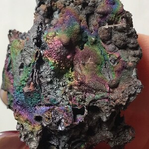 Pu&ograve; includere: Un minerale grigio scuro iridescente con una superficie ruvida e testurizzata. Il minerale ha un bagliore arcobaleno con sfumature di viola, verde e blu.