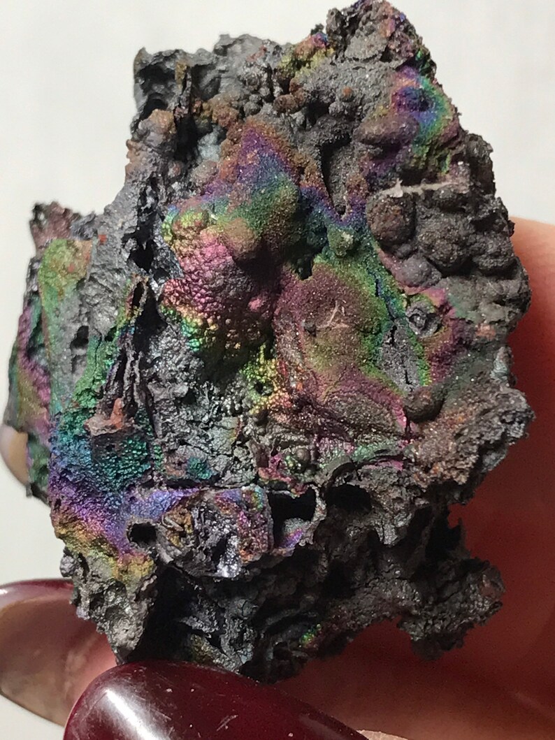 Pu&ograve; includere: Un minerale grigio scuro iridescente con una superficie ruvida e testurizzata. Il minerale ha un bagliore arcobaleno con sfumature di viola, verde e blu.