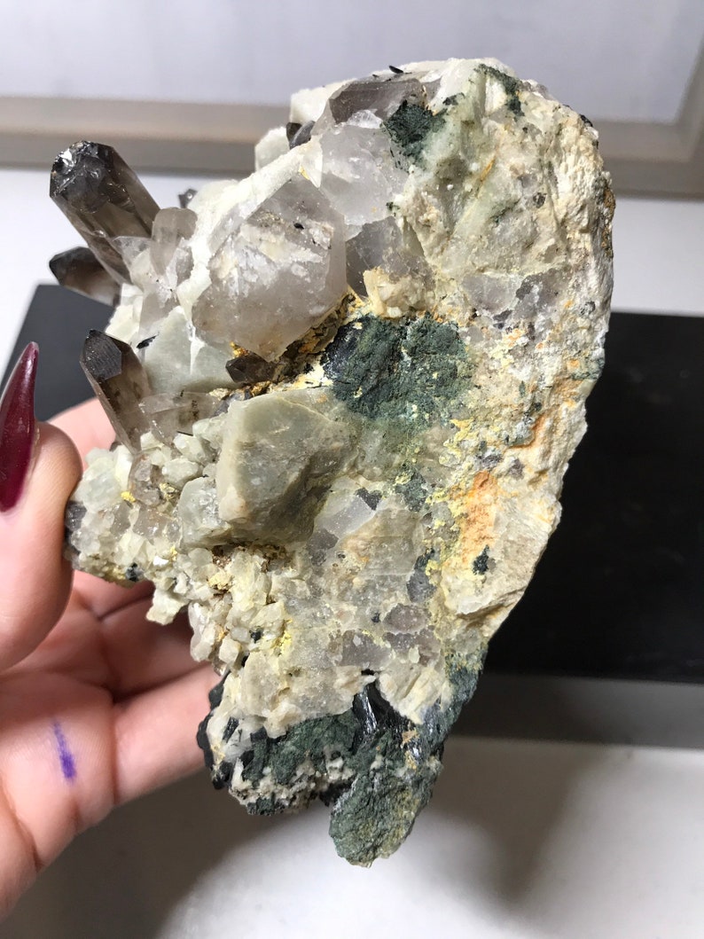 May include: A cluster of smoky quartz crystals embedded in a matrix of white and green rock. The crystals are clear and have a smoky brown color. The matrix is covered in green moss.