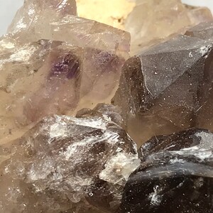May include: Close-up of a cluster of smoky quartz crystals with a hint of purple. The crystals are translucent and have a rough, natural texture.