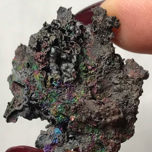 Pu&ograve; includere: Un minerale grigio scuro iridescente con una superficie ruvida e testurizzata. Il minerale ha un'opalescenza metallica con sfumature di verde, blu e viola.