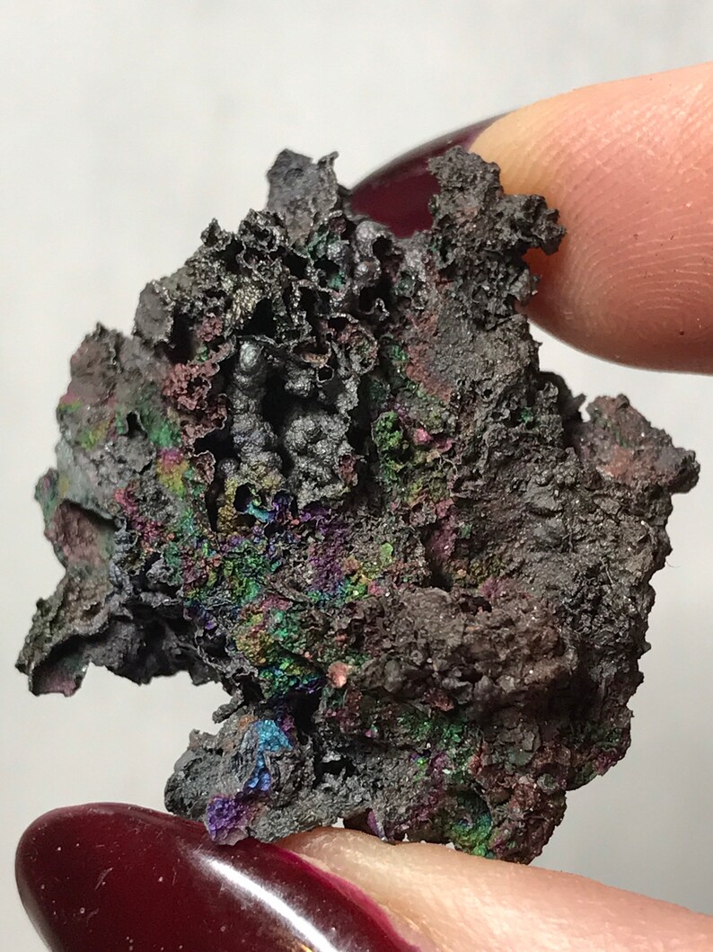 Pu&ograve; includere: Un minerale grigio scuro iridescente con una superficie ruvida e testurizzata. Il minerale ha un'opalescenza metallica con sfumature di verde, blu e viola.