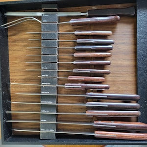 May include: A vintage carving set in a wooden box. The set includes a carving fork and multiple knives with wooden handles and stainless steel blades. The box is black with a wooden interior and a metal clasp.