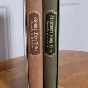 May include: A two-volume set of hardcover books housed in a green slipcase. The left book is tan with a brown spine label titled Grimms' Fairy Tales illustrated by Fritz Kredel. The right book is green with a green label titled Andersen's Fairy Tales.