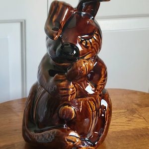 May include: A brown ceramic cookie jar in the shape of a dog, with a glossy finish. The dog is depicted in a seated position, holding a small object. The jar has a lid and is designed with detailed features.