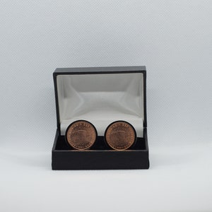 May include: A black gift box with a velvet interior, containing two copper cufflinks. The cufflinks feature a design with the words "Dominion of New Zealand" and a stylized fern leaf.