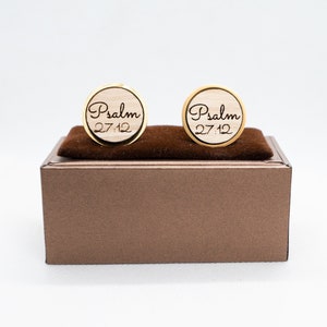 May include: A pair of wooden cufflinks with a gold trim. The cufflinks are round and have the words "Psalm 27:12" engraved on them.