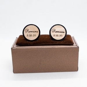 May include: A pair of wooden cufflinks in a brown box. The cufflinks are round and have a black border. The text "Romans 8:38-39" is engraved on the cufflinks.