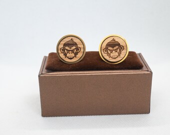 May include: A pair of gold-toned cufflinks with a wood inlay featuring a monkey face design. The cufflinks are displayed in a brown box.