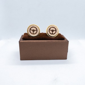 Op de afbeelding: Een paar gouden manchetknopen met een houten inlay van een stuurwielontwerp. De manchetknopen worden gepresenteerd in een bruine doos.