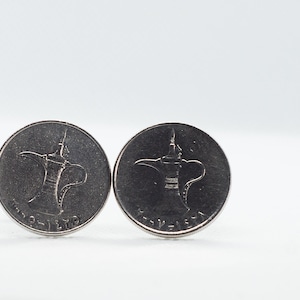 May include: Two silver-colored coins with a design of a traditional Arabic coffee pot on a white background.