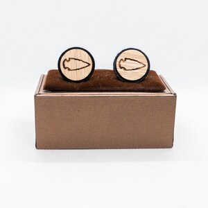 May include: A pair of wooden cufflinks in a brown box. The cufflinks are round and have a black border. Each cufflink features a carved arrowhead design.