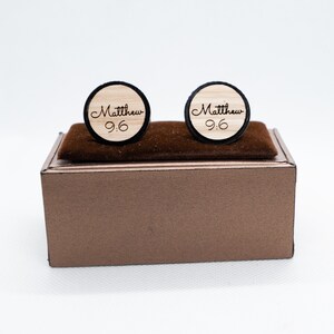 May include: A pair of wooden cufflinks with a black border. The cufflinks are engraved with the name "Matthew" and the number "9:6". The cufflinks are displayed in a brown box.