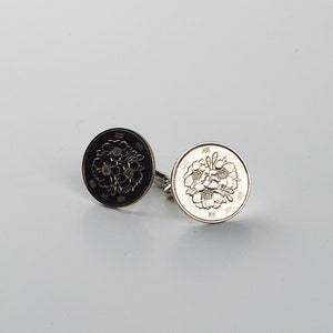 May include: A pair of silver cufflinks featuring a design of five cherry blossoms. The cufflinks are on a white background.