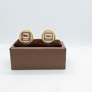 May include: A pair of gold cufflinks with a wooden base. One cufflink has the word "Esc" engraved on it, and the other cufflink has the word "Ctrl" engraved on it.