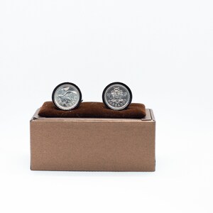 May include: A pair of silver cufflinks featuring coin designs. One cufflink displays a bird and the words "TEN CENTS", while the other shows a coat of arms and the word "BARBADOS". The cufflinks are set against a brown velvet cushion inside a brown box.