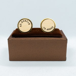 May include: A pair of gold cufflinks with a wooden surface and black engraved numbers. The cufflinks are displayed in a brown box.