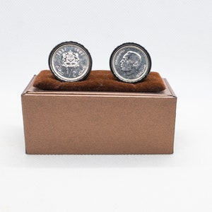 Könnte beinhalten: Ein Paar silberne Manschettenknöpfe in einer braunen Schachtel. Die Manschettenknöpfe zeigen eine silberne Münze mit einem Kronendesign und dem Text "1/2 DH" auf der einen Seite und ein Porträt eines Mannes auf der anderen.