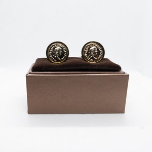 Op de afbeelding: Een paar gouden manchetknopen met een muntontwerp, gepresenteerd in een bruine fluwelen doos.