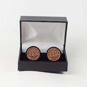 Può includere: Un paio di gemelli in una scatola nera. I gemelli sono realizzati con monete canadesi da 1 centesimo del 1963. Le monete presentano un design a foglia d'acero e il testo "1 CENT 1963 CANADA".