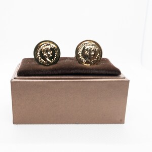 Op de afbeelding: Een paar gouden manchetknopen in een bruine fluwelen doos. De manchetknopen hebben een reliëf van een gezicht.