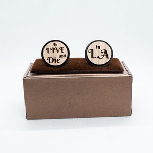 May include: A pair of wooden cufflinks in a brown box. The cufflinks are round and have black edges. One cufflink says "To Live and Die" and the other says "in L.A."