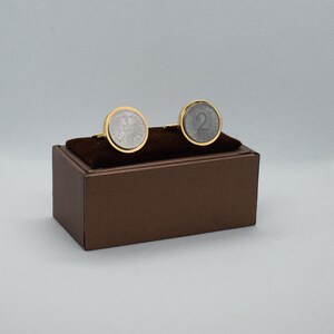 May include: A pair of cufflinks in a brown box. The cufflinks are made of gold-plated metal and feature a silver coin design. One cufflink features a German eagle, and the other features the number "2".