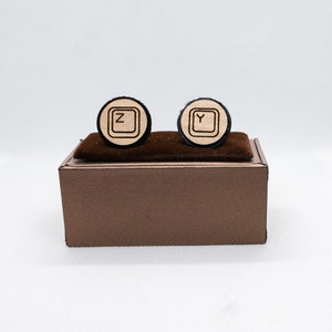 May include: A pair of wooden cufflinks in a brown box. The cufflinks are round and have a black border. The cufflinks are engraved with the letters "Z" and "Y" in a keyboard style font.