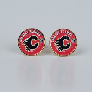 Puede incluir: Dos gemelos de oro con logotipos de los Calgary Flames en rojo, blanco y negro. Los logotipos representan una llama estilizada con una "C" en su interior.