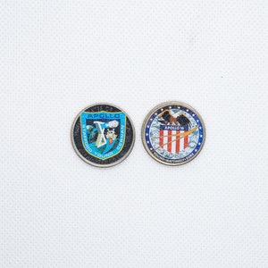 Peut inclure: Deux pièces commémoratives rondes de couleur argent avec des motifs bleus et blancs. La pièce de gauche porte le texte "Apollo 10" et les noms "Stafford, Young, Cernan". La pièce de droite porte le texte "Apollo 16" et les noms "Young, Mattingly, Duke".