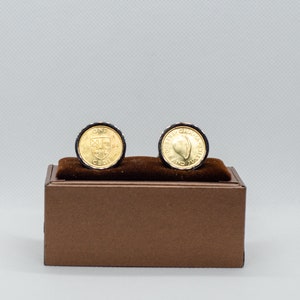 May include: Two gold cufflinks with black borders, featuring a design of a shield with a cross and the words "ONE CEDI" on one cufflink and a design of a leaf with the words "REPUBLIC OF GHANA FREEDOM AND JUSTICE" on the other cufflink. The cufflinks are displayed on a brown box.