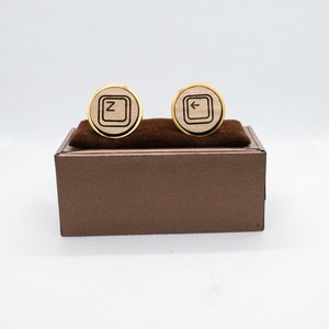 May include: A pair of round cufflinks with a gold-colored border. One cufflink features the letter "Z" inside a square, and the other has a left arrow symbol. The cufflinks are made of light-colored wood and are displayed on a brown box.