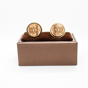 Può includere: Un paio di gemelli rotondi con un design geometrico a tartaruga inciso nel legno chiaro. I gemelli hanno un bordo color oro e sono esposti su un cuscino di velluto marrone all'interno di una scatola marrone. Lo sfondo è bianco.