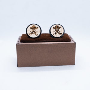 Può includere: Un paio di gemelli in legno in una scatola marrone. I gemelli sono rotondi e presentano un design di teschio e ossa incrociate con un cappello da pirata.