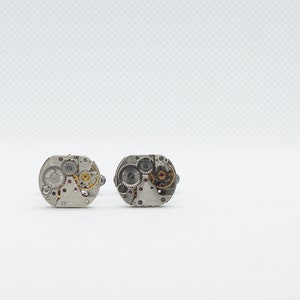 May include: A pair of silver cufflinks with intricate clockwork designs. The cufflinks are made of metal and have a circular shape.