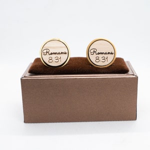 May include: A pair of gold cufflinks with a wooden inlay. The cufflinks are engraved with the text "Romans 8:31".