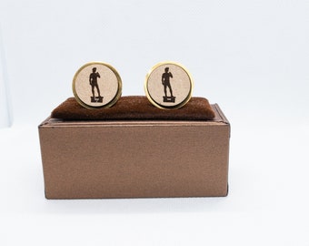 May include: A pair of gold cufflinks with a beige background featuring a silhouette of a statue. The cufflinks are displayed in a brown box.