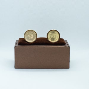 Könnte beinhalten: Ein Paar goldene Manschettenknöpfe in einer braunen Schachtel. Die Manschettenknöpfe sind rund und zeigen ein Design mit einem Vogel und einem Wappen.
