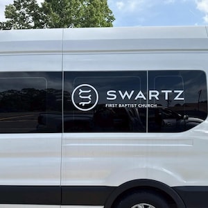 May include: A white passenger van with black trim and tinted windows. The van has the name "SWARTZ" in large white letters, along with the words "FIRST BAPTIST CHURCH" below. A circular logo is on the left side of the text.