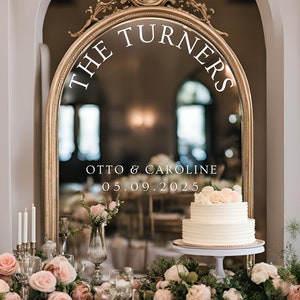 May include: A wedding cake with white frosting and pink flowers sits on a white cake stand in front of a large ornate mirror. The mirror reflects the text "The Turners Otto & Caroline 05.09.2025".