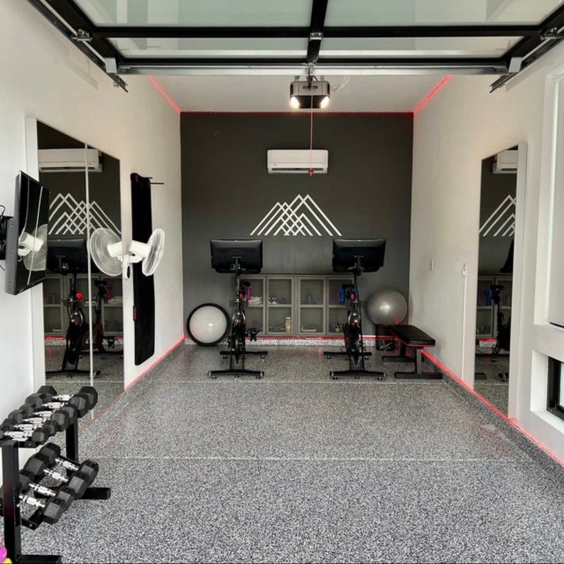 Puede incluir: Un gimnasio en casa con dos bicicletas est&aacute;ticas, un juego de pesas y un ventilador. Las paredes son blancas y grises, y el suelo es gris moteado. Hay un espejo en la pared izquierda y una ventana en la pared derecha.