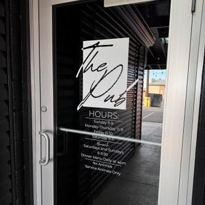 May include: A glass door with a white sign displaying the name "The Dub" in cursive lettering. The sign also lists opening hours, including brunch times on Saturdays and Sundays. The door has a silver handle and is set in a black frame.
